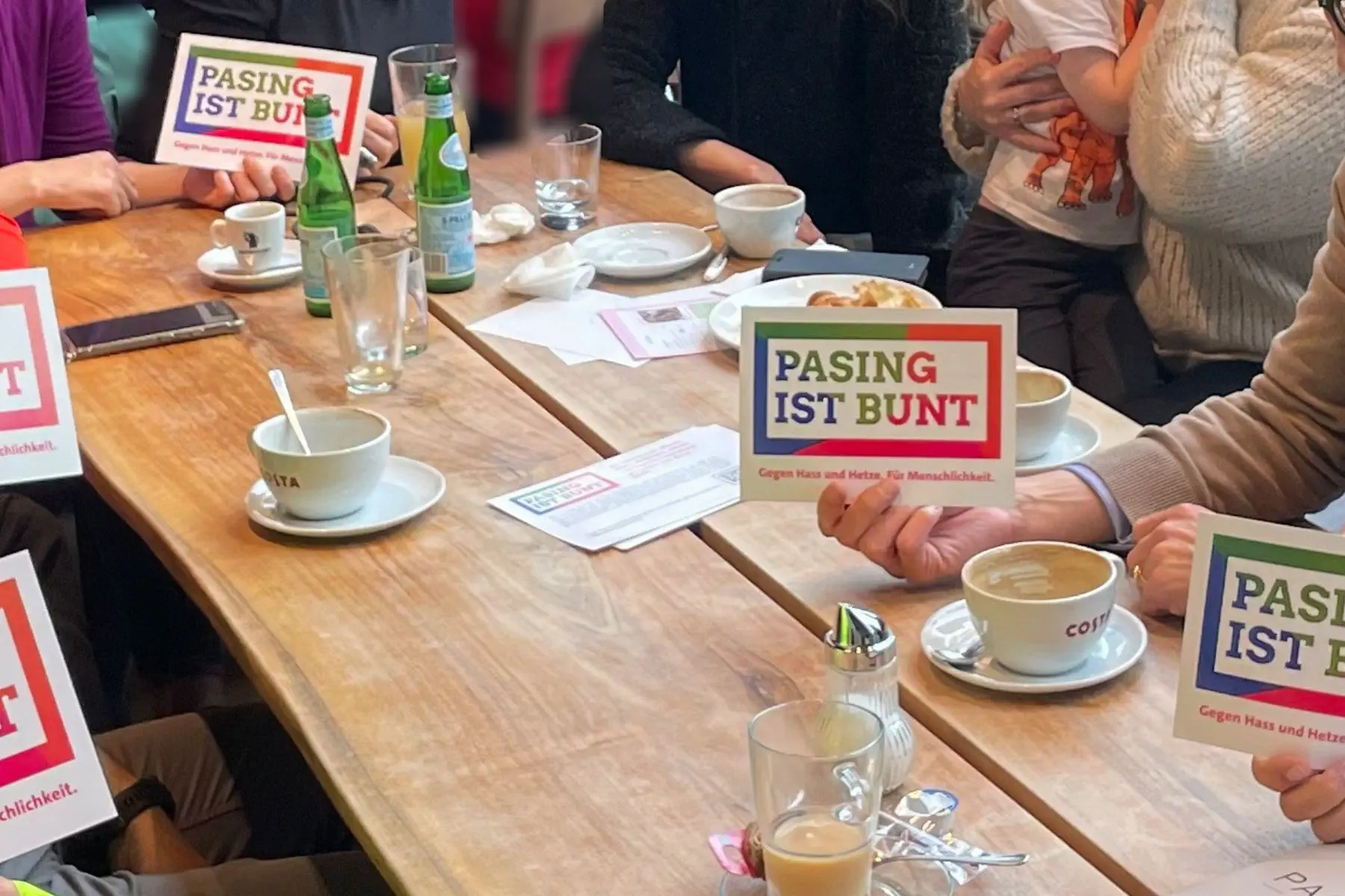 Tisch mit Kaffeetassen und vielen Händen, die Pasing ist bunt Schilder halten